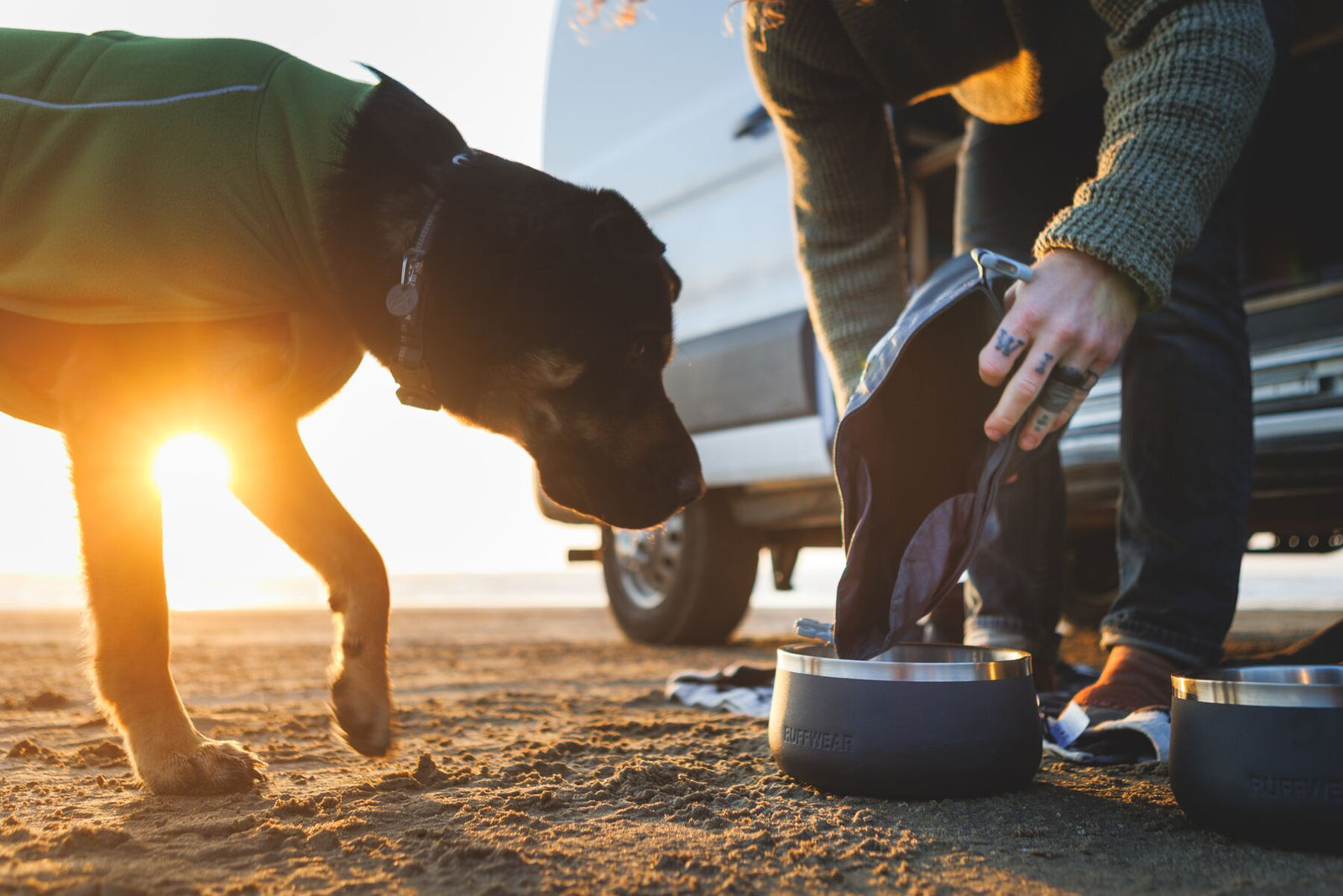 Person crouching on a sandy beach at golden hour pouring kibble from the Ruffwear Kibble Kaddie into a Ruffwear Basecamp Bowl, with a black dog in a green Ruffwear coat watching eagerly, a campervan visible behind them